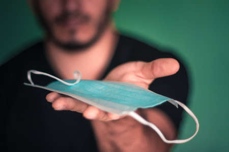 A young adult man with a black t-shirt showing a light blue disposable mask with folds and white strips on the palm of his hand on a sea green background alluding to the theme of the COVID-19 pandemic known as coronavirusの写真素材