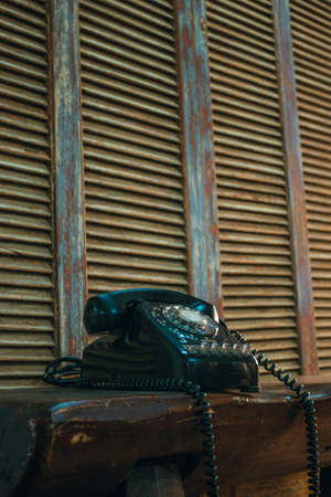 old obsolete black telephone on a wooden table in the middle of a room with a wooden shutter in the...
