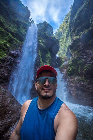 adult tourist man wearing cap and sunglasses with two beautiful waterfalls falling into the riverbed in the middle of the tropical jungle of Costa Ricaの写真素材