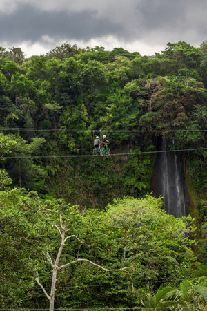vertical shot couple of hikers with harness and helmet riding zipline bicycles surrounded by vegetation and a beautiful waterfall in Costa Ricaの写真素材