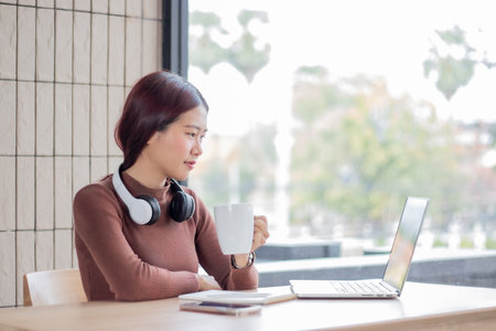 Young Asian woman uses a computer, wearing headphones and using a notebook for an online video...