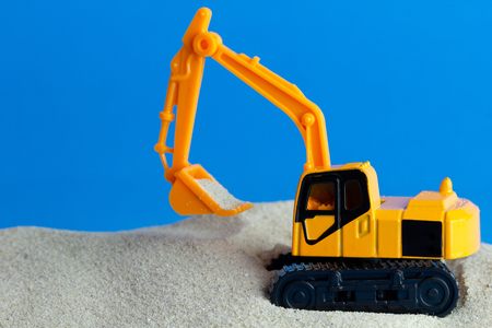 Yellow toy backhoe is digging sand on the blue background. Concept of construction.の写真素材