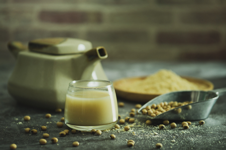 Soymilk in the glass and the kettle is placed beside. Soybean powder is crushed in a wooden dish and has scattered soy beans on the table in morning light.の写真素材