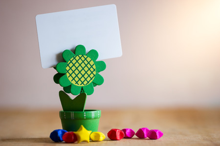 Clip holder card stand green sunflower shaped and multicolored paper stars on wooden table. White plastic blank card in morning sunlight.の写真素材