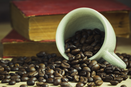 Coffee bean in tilted ceramic cup and old book laid behind on wooden table with morning light.の写真素材