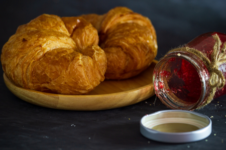 Croissant on wooden dish and strawberry jam bottles in dark background.の写真素材