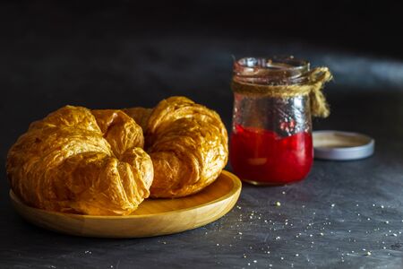 Croissant on wooden dish and strawberry jam bottles in dark background.の写真素材