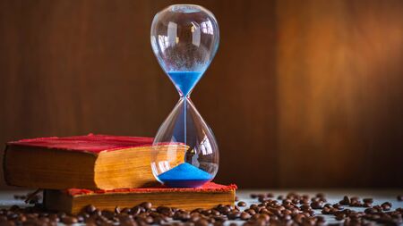 Sand clock on the old book and coffee seed on wooden table. Copy space for text. The concept of time...