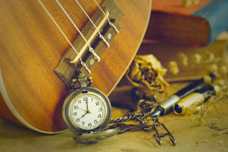 An antique pocket watch leaned against a ukulele and old book with vintage map and brass pen placed on wooden table. closeup and copy space for text. The concept of memories or things in the past.の写真素材