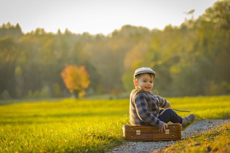 Happy little boy playing on road at sunset. He standing in the field and holding in hands suitcase and balloons.の写真素材