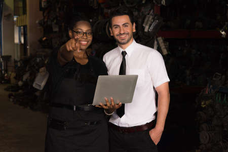 Diversity of two people, black African worker woman in a warehouse holding computer-notebook and pointing to car part in a factory while caucasian salesman business manager looking at her pointingの写真素材