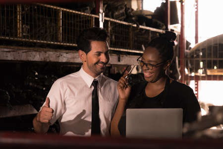 Diversity of two people, caucasian business manager give a thumb up to black African worker woman in a warehouse while she smiling and holding computer-notebook in factoryの写真素材