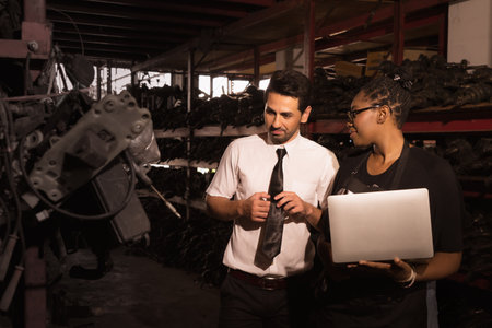 Diversity of two people, a caucasian business manager looking at car part in factory-warehouse and serious talking with black African worker woman who holding laptop-notebookの写真素材