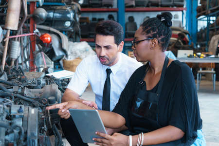 Diversity of two people, black African worker woman explain about engine to caucasian business manager in factory-warehouseの写真素材
