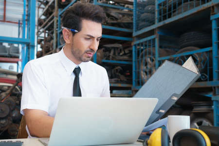 Caucasian business manager sitting and reading report in front of laptop-notebook in factory-warehouseの写真素材