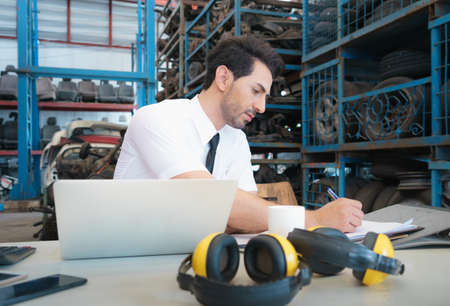 Caucasian business manager sitting and writing report in front of laptop-notebook in factory-warehouseの写真素材