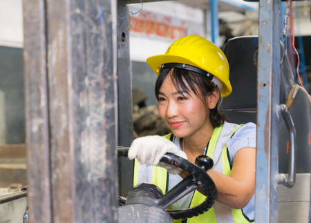 Cute Asian engineer worker women looking left side while driving forklift in factory-warehouseの写真素材