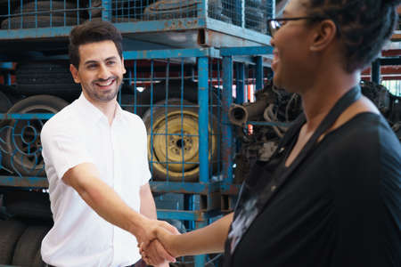 Man stand and shakehand with woman after deal succeed. Diversity of two people, caucasian business manager work with African worker woman in factory-warehouseの写真素材