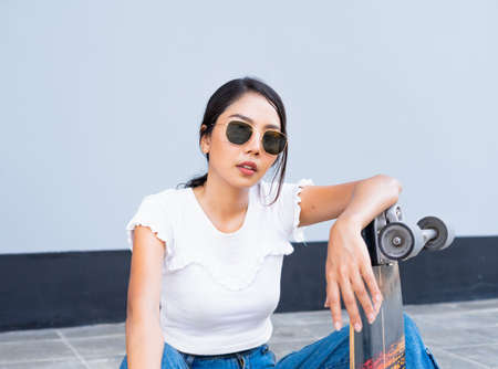 Half body of Asian cute girl or woman sit on street, place skateboard in vertical, and act smart in front of cool wall with copy space in daylight time, summer holidayの写真素材