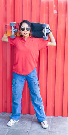 Asian cute girl or woman stand, hold skateboard above shoulder, looking straight, and act smart at red cool wall in summer holidayの写真素材