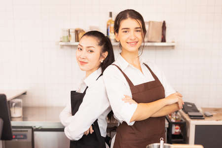 Beautiful caucasian and Asian barista women stand with crossed arms to welcome customers at coffee...