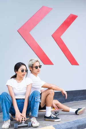Asian handsome man and cute woman sitting on skateboards, wear sunglasses, look at right side in...