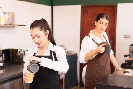 Asian barista women fill milk into takeaway hot coffee cup while caucasian barista woman looks at her to prepare coffee for customer in cafe coffee shop. Barista work at coffee bar and food service businessの写真素材