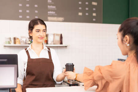Beautiful caucasian barista woman give takeaway coffee cup to customer and customer receiving coffee cup, focusing at face of barista woman. Barista work at food and coffee service business.の写真素材