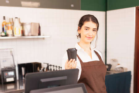 Beautiful caucasian barista woman show takeaway coffee cup in one hand at coffee bar. Barista work at coffee bar and food service businessの写真素材