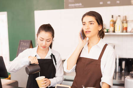 Caucasian barista woman confirms online orders with customer via mobile, Asian barista woman fills white milk into takeaway coffee cup at cafe bar. Barista work at coffee bar and food service businessの写真素材