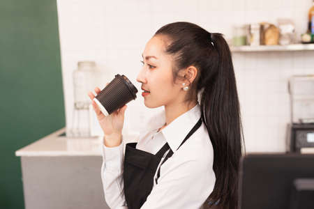 Left angle of beautiful Asian barista women enjoy hot black coffee at coffee bar in cafe coffee...