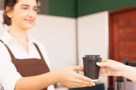 Right side of takeaway coffee cup in two hands of customer and beautiful caucasian barista woman who...