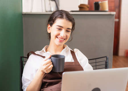 Beautiful caucasian barista woman sits relaxing to drink hot coffee in breaking time and happy to use laptop at coffee shop. Barista work at coffee bar and food service businessの写真素材