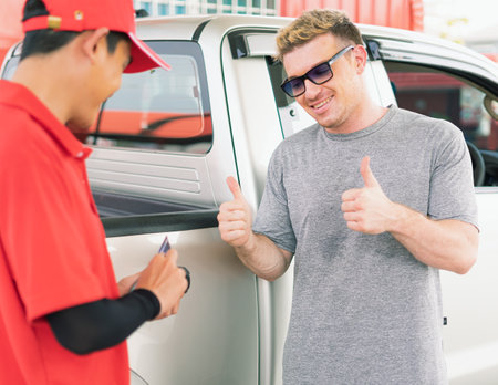 White caucasian car owner give thumbs up for credit card payment succeed by Asian gas station worker man and after filling high energy power fuel into auto car tank in petrol station, commercial service for benzine, diesel, gasohol, gasoline.の写真素材