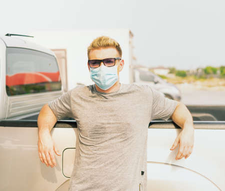 White caucasian wear virus facemask and stand with placing arms on car while waiting for filling high energy power fuel into auto car tank in petrol station, commercial service for benzine, diesel, gasohol, gasoline.の写真素材