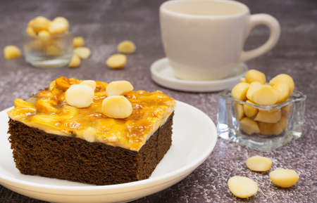 Toffee chocolate cake slice on plate with macadamia seeds in small bowl and coffee cup on stone background for dessert break. Toffee cake made from bake, dark chocolate, macadamia, nut, caramelの写真素材