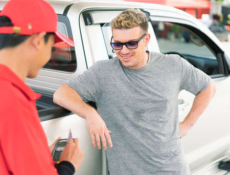 White caucasian car owner standing and looking at his credit card payment by Asian gas station worker man after filling high energy power fuel into auto car tank in petrol station, commercial service for benzine, diesel, gasohol, gasoline.の写真素材
