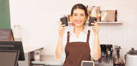 Beautiful caucasian barista woman happy to hold two takeaway hot coffee cups at the coffee bar in cafe coffee shop. Barista work at coffee bar and food service businessの写真素材