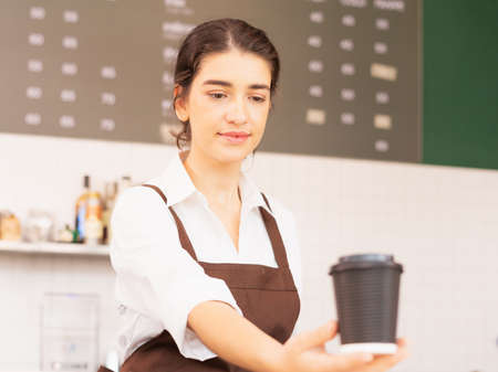 Right side of beautiful caucasian barista woman shows takeaway coffee cup in one hand forward and looking to coffee cup at coffee bar. Barista work at coffee bar and food service businessの写真素材