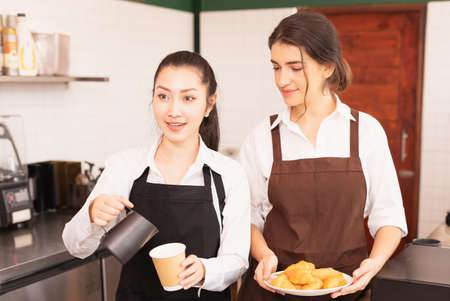 Focusing on half-body angle of Asian barista women stop filling milk into takeaway hot coffee cup while talking with customer in cafe coffee shop. Barista work at coffee bar and food service businessの写真素材
