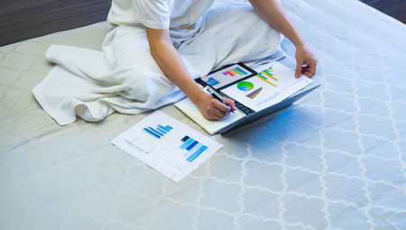 asian business man work with chart or graph report and note on notebook on bed, worker sits and works business on chart or graph for data analytics on bed at homeの写真素材