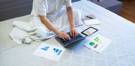 asian business man type keyboard on laptop on bed, worker sits and works business on chart or graph for data analytics on bed at homeの写真素材