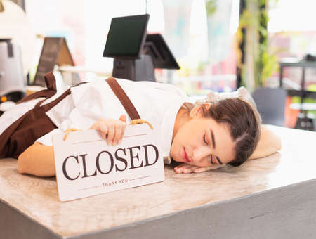 Higher right side angle of beautiful caucasian barista woman relax sleeping with show closed shop signs on coffee bar. Barista work at coffee bar and food service businessの写真素材