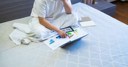 asian business man work online meeting with chart or graph reports on bed, worker sits and works business on chart or graph for data analytics on bed at homeの写真素材