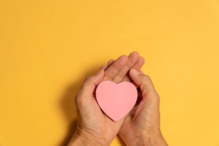 Female hands holding a pink heart on a yellow background with copy spaceの写真素材