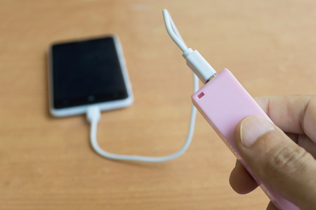 businessman hand holding a pink powerbank with smartphone on wood deskの写真素材