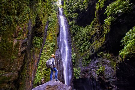 Young man with straw hat relaxes looking at the Sekumpul waterfalls in jungles on Bali island,...