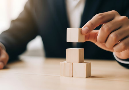 A hand in a dark suit places a blank wooden cube atop a stack of three on a light wood surface.の素材