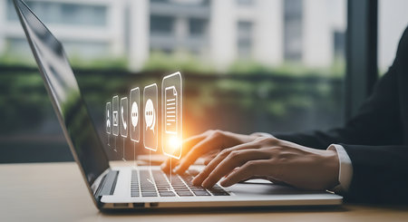 Hands typing on a laptop with glowing digital icons for communication and documents appearing above the keyboard.の素材