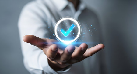 A hand holds a glowing blue checkmark within a circular outline, surrounded by shimmering digital particles against a dark background.の素材